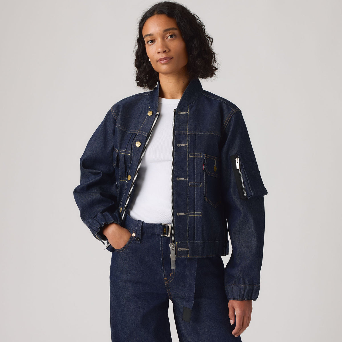 A model wearing a dark indigo denim bomber jacket with contrast stitching, zippered pockets, and a belted waist, styled over a white t-shirt and matching dark denim jeans against a light gray studio background.