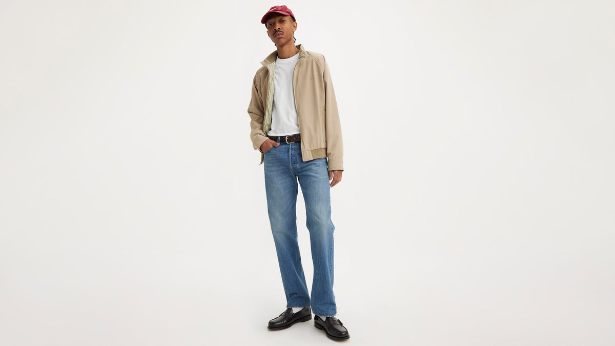 Full-length front view of man in beige jacket, mid-blue Levi’s 501 jeans, & red cap
