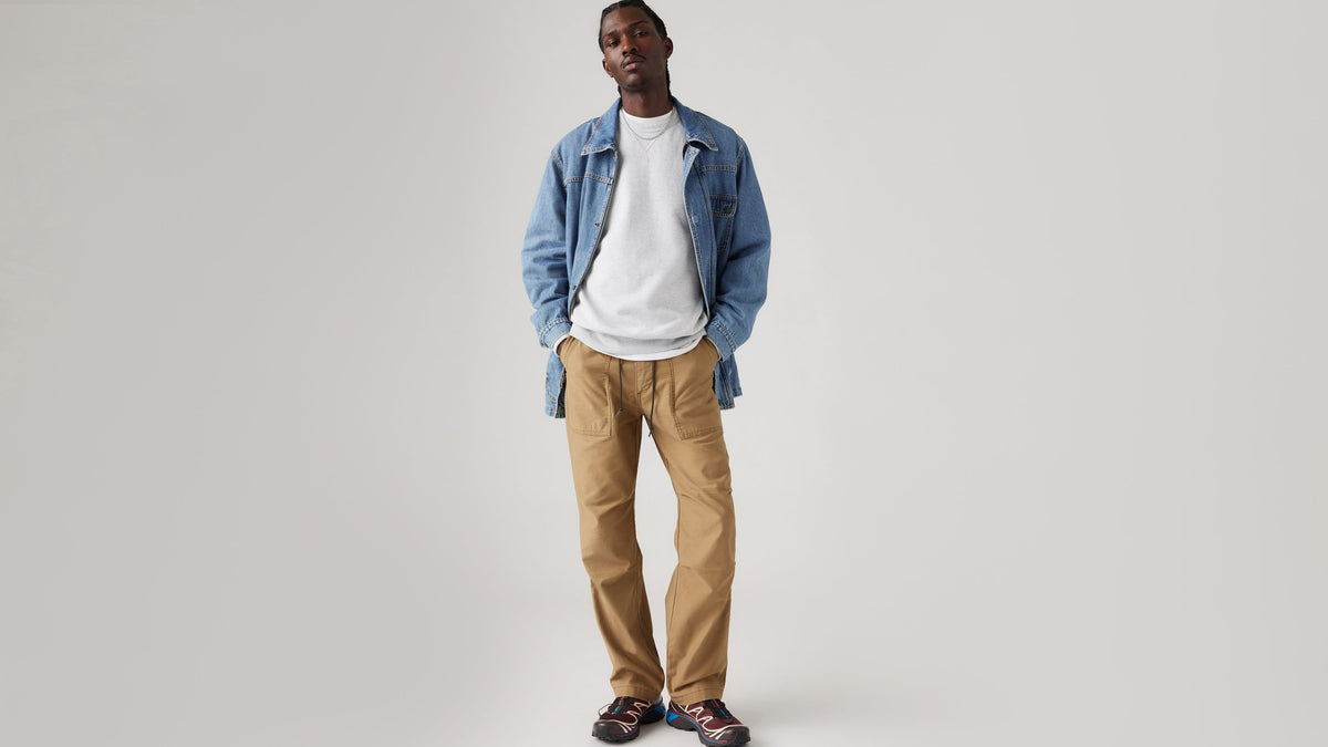 Front view of a man wearing khaki Loose Surplus Pants, a denim jacket, & sneakers