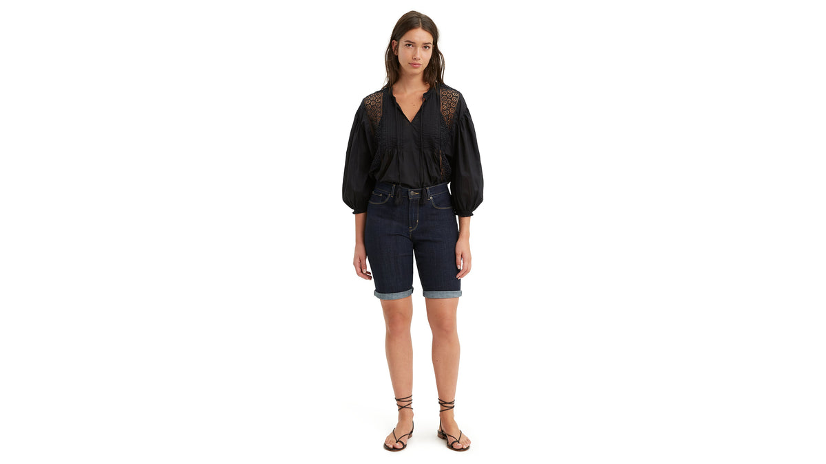 Woman in dark wash Levi’s knee shorts with a black blouse & strappy sandals