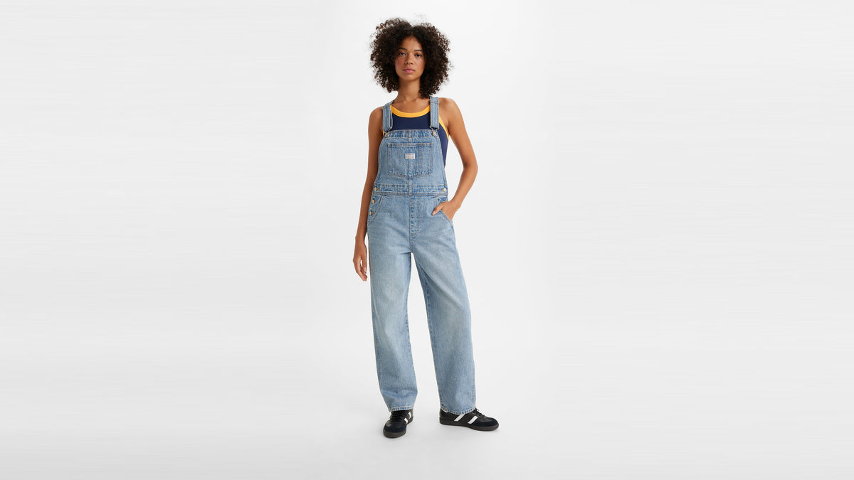Full-body view of a woman in light blue vintage overalls with tank top & black sneakers