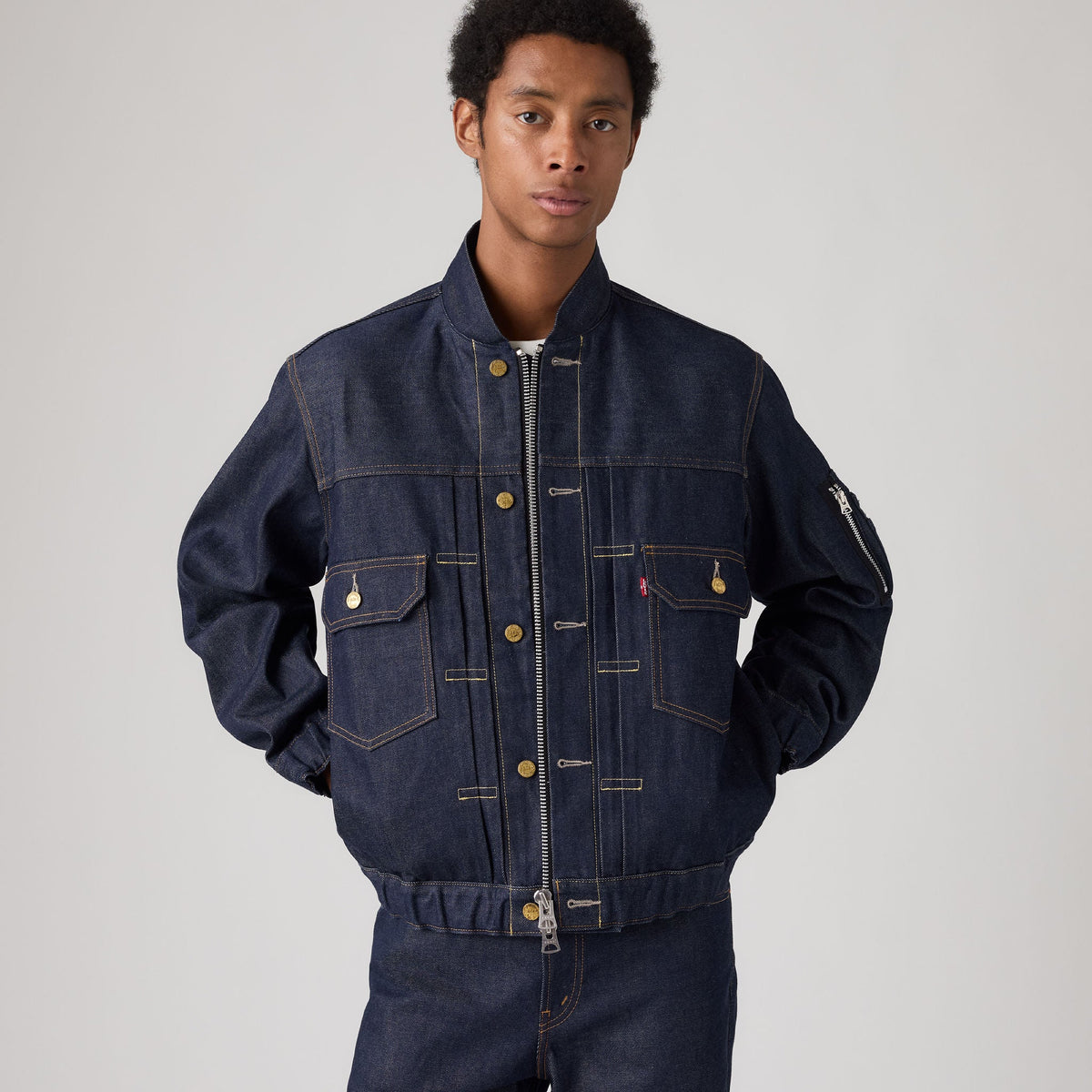 A man wearing a dark indigo denim jacket with a front zipper, button snap closure, and chest pockets, paired with matching dark denim pants, shot against a neutral light gray studio background.